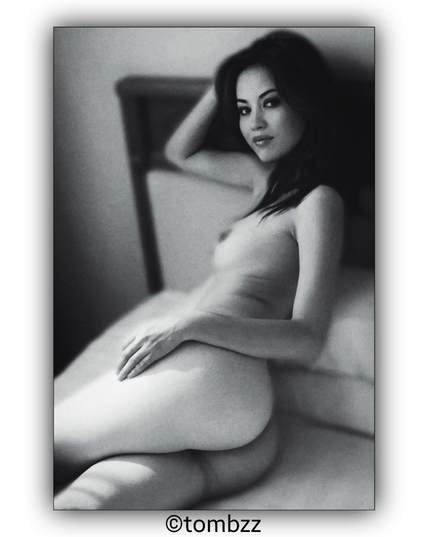 A nude portrait of a young woman with long, dark hair. The model is lying on her side, leaning against the headboard of the bed, with one hand on her hip. She gazes directly into the camera. The image is in black and white.