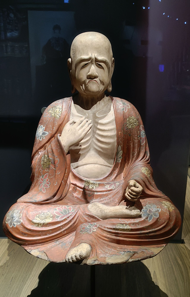 Photographie numérique couleur de format portrait, avec une statue de moine dans une vitrine, où on voit l'ombre de l'objet sous son socle et le fond en bois clair. Il y a des reflets d'éclairages dans la vitre.
Le moine est assis en tailleur avec la jambe droite repliée sur la gauche. Il porte un vêtement de couleur saumon pâle avec des motifs de fleurs grises et jaunes. 
Son visage exprime de la tristesse, voire de la colère : froncils froncés, bouche serrée et mécontente. Le vêtement est ouvert sur son torse où ses côtes sont saillantes. Sa main droite est posée sur son torse, la droite est repliée le long de son corps.