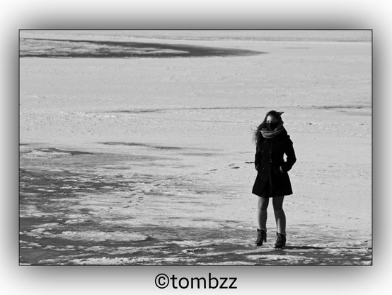 A black and white photograph of a girl standing on ice. She is wearing a dark coat and boots. The background shows a vast, frozen surface with patches of snow and ice, giving the scene a stark and cold atmosphere. The girl appears to be alone in this expansive, icy landscape.