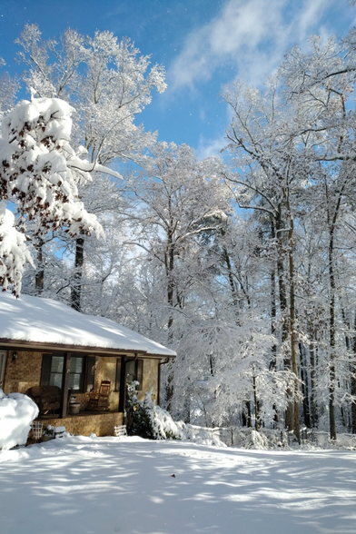 A sunny morning after snow. Blue sky with light clouds, snow covered trees, and a ranch house on the left with the rooftop covered with snow, and the bottom is snow covered ground.