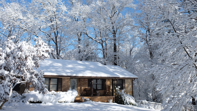 A sunny morning after snow. Blue sky with light clouds, snow covered trees, and a ranch house in the middle with the rooftop covered with snow, and the bottom is snow covered ground.