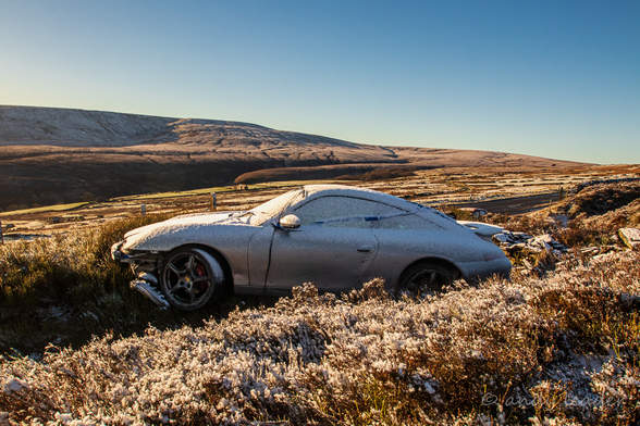 A rather fancy, powerful car rests amongst the heather moors covered in frost after coming off the road. Blue sky and frosty hilss the backdrop.