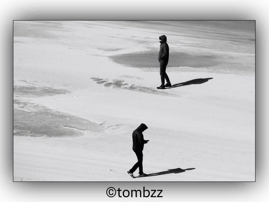 A black and white photograph of a winter beach. Two men are walking separately, each casting a distinct shadow on the sand. One man is looking at a device in his hands while the other has his hands in his pockets. The image captures the stark contrast between the figures and their surroundings.