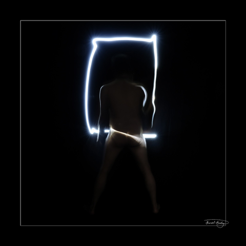 A very dark square image of a standing nude male from behind who is painting a frame with a flashlight in front of him. One ray of light goes over his bottom.