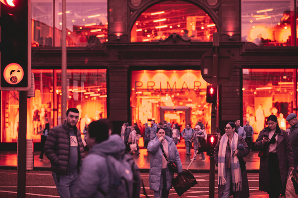 This image captures a bustling city street bathed in deep red and orange hues through infrared photography, evoking a surreal, dystopian atmosphere. In the foreground, pedestrians in heavy winter coats move across the street, their faces blurred and indifferent, as if trapped in routine. A glowing red traffic light dominates the scene, its stop signal commanding control like an omniscient eye. Behind them, a grand department store looms, its large windows glowing with intense orange light. Mannequins stand stiff and faceless inside, mirroring the pedestrians outside. Bright reflections streak across the windows, distorting reality and adding tension. The fiery palette transforms the street into a symbolic space of uniformity and quiet resignation. The pedestrians appear small and anonymous, their individuality dwarfed by the overwhelming architecture and oppressive red glow, evoking themes of conformity and control in an Orwellian urban world.