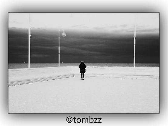 A black and white photograph depicting a snowy pier. A woman is standing on the pier, facing away from the camera. The sky is filled with dark clouds, and in the distant background, ships can be seen anchored offshore. The scene is framed by tall, slender lamp posts.