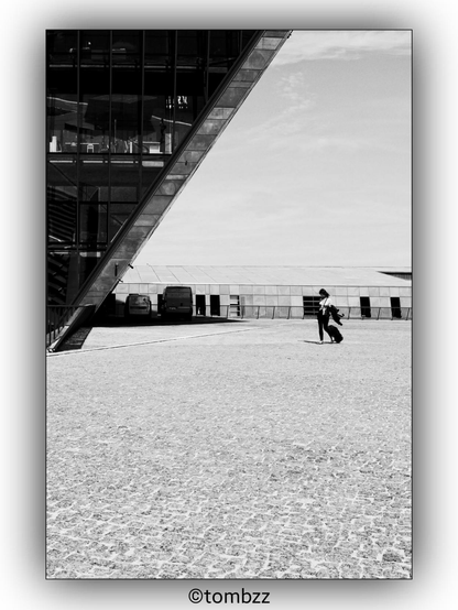 A black and white photograph showing a girl walking across a large, cobblestone square while pulling a suitcase. On the left side of the image, there is a prominent modern building with a diagonal structure made of glass and metal. In the background, there are additional buildings and a clear sky, adding depth to the scene.