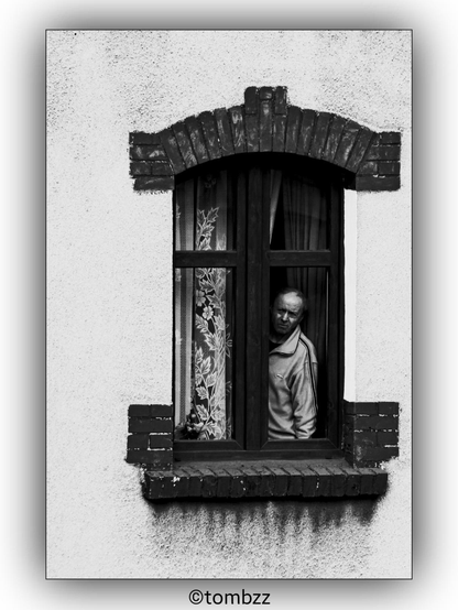 A black and white photograph of a man looking out of a window in an old tenement building. The window features decorative elements such as a brick arch above it and brick details on the sides and bottom. The man appears intrigued or surprised by something outside. The window has curtains with floral patterns, adding a touch of vintage charm to the scene.