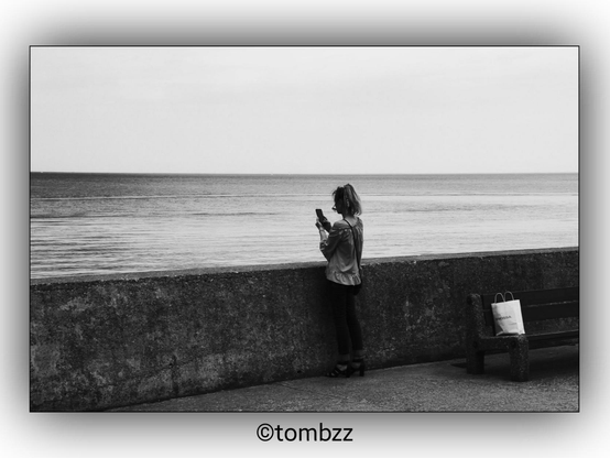 A black and white photograph showing a girl standing on a seaside promenade, holding a smartphone in her hands. Beside her, there is a bench with a paper bag placed on it. The sea can be seen in the background, adding a serene atmosphere to the scene.