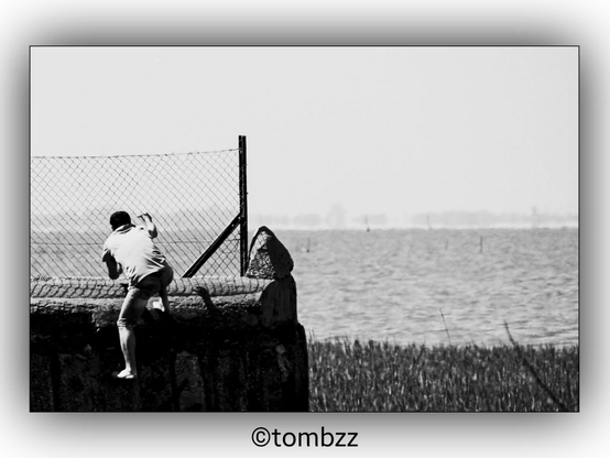 A photograph showing a man climbing over a wall and a fence. In the background, there is a reed bed and the waters of a lagoon, adding a natural and scenic element to the image.