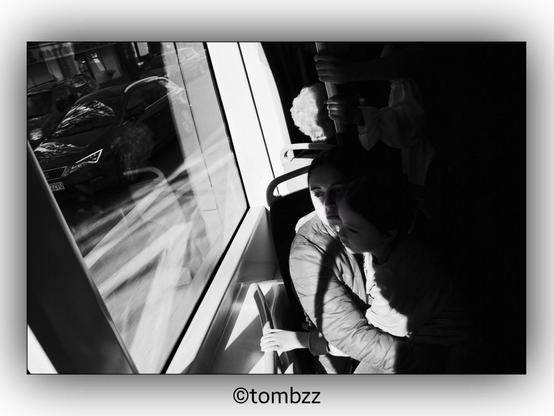 A black and white photograph showing a mother and her daughter sitting together on a tram. The mother is holding the daughter on her lap, and both are looking out of the window. The daughter is holding onto a pole. The image captures a beautiful play of light and shadows, with reflections of the street and a car visible on the tram window. The scene highlights a quiet, intimate moment between the mother and daughter, enhanced by the interplay of light and reflections.