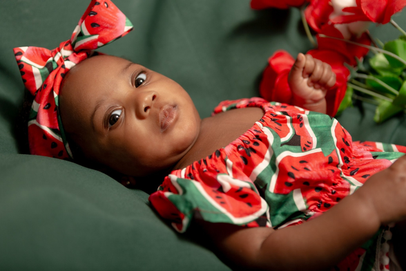 #Newborn #Photography of beautiful black newborn looking into the camera over her right shoulder laying (profile) down on a dark green background and dressed with a short sleeve romper decorated with lots of big watermelon slices on a dark green background.