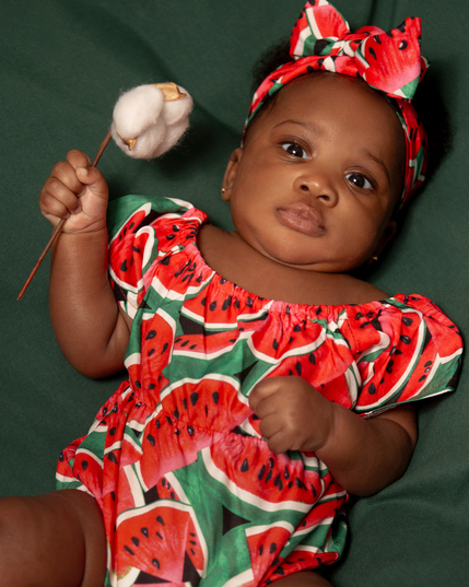 #Newborn #Photography of beautiful black newborn babygirl looking straight into the camera, laying on her back, diagonally from right to left, on a green background, dressed with a short sleeve romper decorated with  big watermelon slices on a green background and holding a cotton flower in her right hand.