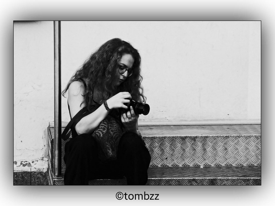 A black and white photograph of a woman with long, curly hair sitting on a metal step while holding a camera. She is focused intently on the camera, possibly adjusting settings or reviewing a photo. The background features a plain wall and part of a metal railing, giving the image a minimalist feel.