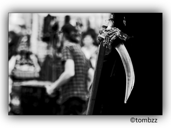 A street photo showing a person dressed as Death holding a scythe in the foreground. In the background, there are people walking and going about their day. The image captures the contrast between the costume and the everyday street scene.