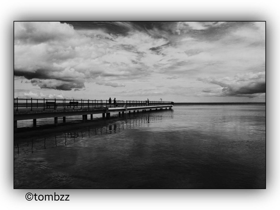 A black and white photograph of a pier extending into a calm body of water. The pier has railings and benches along its length. Two silhouetted figures are visible walking on the pier. The sky is filled with dramatic clouds, adding depth and contrast to the scene. The water reflects the pier and the sky, creating a serene and contemplative atmosphere.