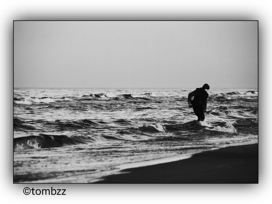 A black and white photograph showing a person wading through the waves of the sea, possibly searching for amber. The sea is slightly rough with visible waves. The person is dressed in dark clothing, which contrasts with the lighter background of the sea and sky. The photo has a timeless and dramatic feel, capturing a moment of quiet determination.