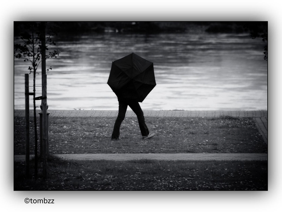 A black and white photograph of a person walking along a riverside path holding a black umbrella. The background features a calm river. The person is wearing dark clothing and appears to be walking on a grassy area next to a paved path. The scene captures a serene moment with a touch of mystery.