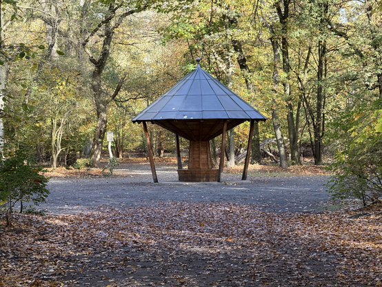 A small structure stands in a wooded area, the structure has a round bench at the bottom, with the middle leading up to a much bigger pointed roof. The roof is held up by big wooden boards.