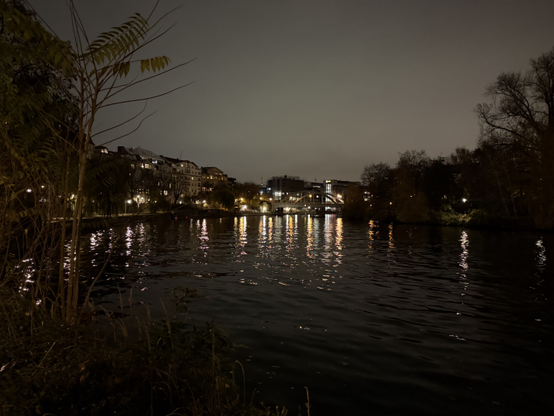 The river Spree with trees and lights lining the left side, and mostly trees and very few lights lining the right side. In the distance there is a bridge, and many more lights, most of which are reflected in the water of the river.