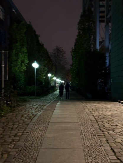 A path made of cobblestone, tiles and small stones bathed in an off-screen light, and then suddenly transitioning into darkness, with the shadow of a building blocking the aforementioned light.

The dark area of the path is dimly lit by green street lamps that are lining the left side of the path.