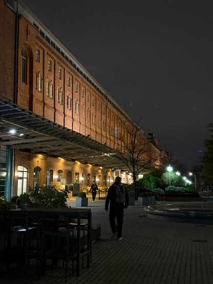 A square with a brightly lit red brick building. The building has outdoor seating at street level, this outdoor seating is covered by an overhang, over which the brick is especially accentuated by light.