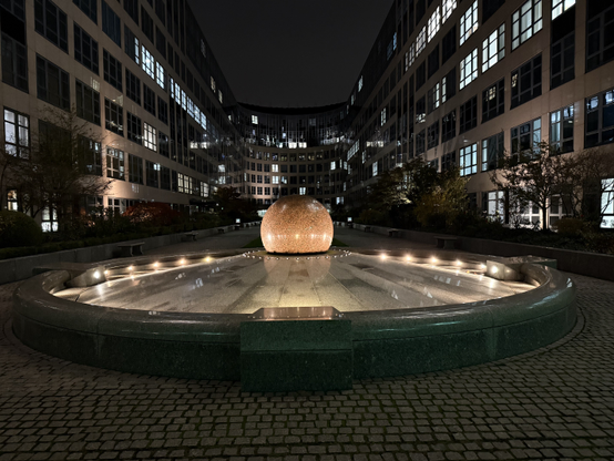 A circular fountain on a cobblestone square. The fountain is turned off, and the inside of the circular rim is lined with lights. The floor of the fountain sloped up to the middle, where there is a big marble. Both the floor and the marble reflect lights.