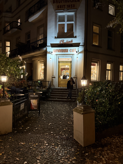 The front of a corner building and the path leading up to it.

The corner is blunted and a door, window and sign are on the blunted corner. This corner is brightly lit, highlighting the golden brown wooden door and the top sign says "Buchwald"' in an old-fashioned cursive font with many flourishes. The bottom sign says "Konditorei Cafe" in a more conventional font.

The entrance path is lit by small latterns.

Picture taken in Moabit, on the Spree.