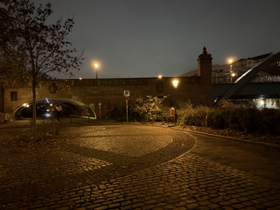 Picture of a cul-de-sac, with orange street lights providing all the light, with a lot of it reflected from the cobblestones. Behind the cul-de-sac is a bridge that only allows pedestrians and bicyclists through, the bridge continues to cross the Spree.

Picture taken in Moabit, following the Spree.