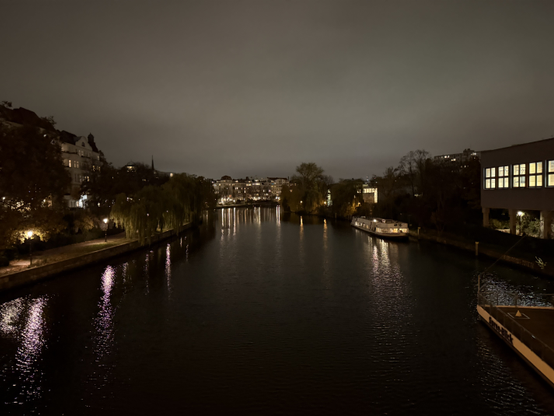 The Spree at night as seen from a bridge, a lower path with trees is seen on the left side, while buildings and a boat are on the right.

The left path has street lights, and a building peaking through the trees, while the building and boat on the right have their own lighting on the inside.

In the centre, there are buildings and lights in the distance.

All lights are reflected in the river.

Photo taken in Moabit.