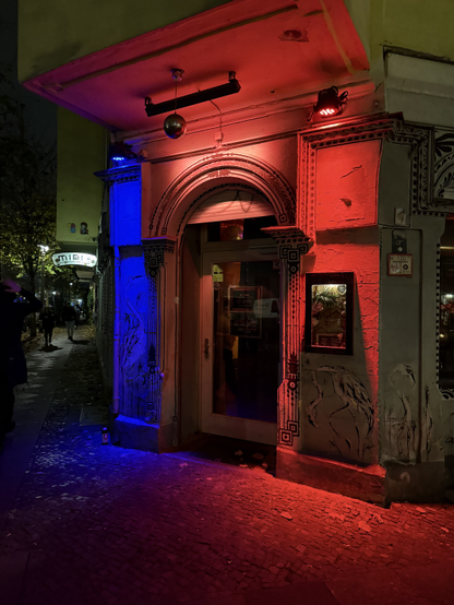 A corner entrance door bathed in colourful light.

The right and centre are bathed in red light coming from lamps hanging above the door, while the left is bathed in blue/violet light from a lamp hanging above its section.

Photo taken in Friedrichshain.