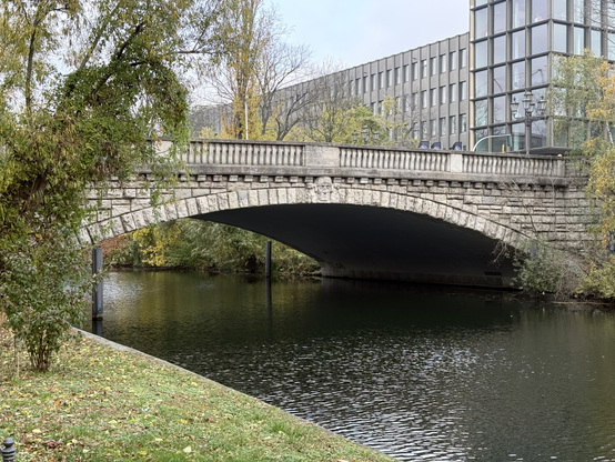 Picture of a grey bridge, taken from a grass field perpendicular, and lower of it. The bridge is made of stone, and has a face in the middle of it.

The photo was taken in Charlottenburg.