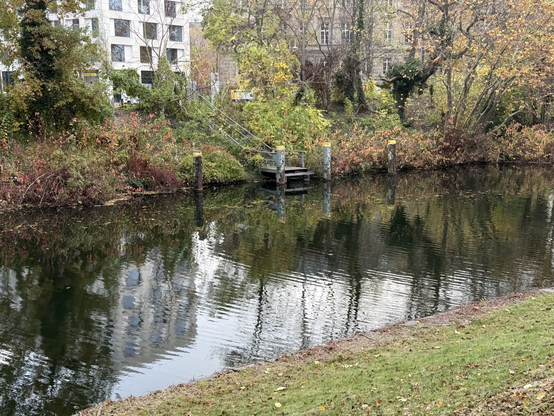 A picture of a platform on the shore of a canal. Stairs lead down to the platform from street level. The platform and the stairs are surrounded by trees, shrubs and other greenery, some of it exchanging their summer uniform for an autumn one.

Picture taken in Charlottenburg.