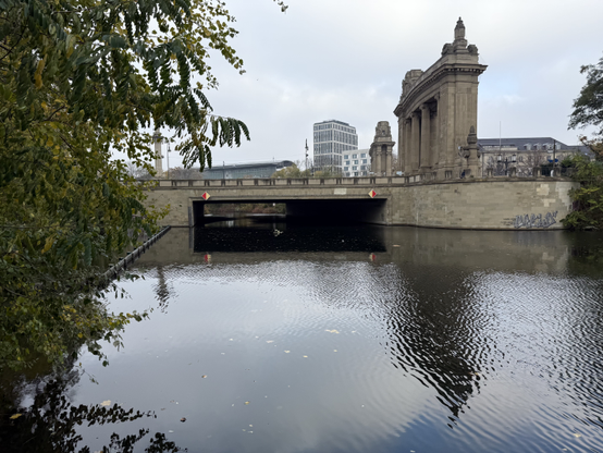 A bridge on a cloudy day. At the bottom is water, reflecting the light from the sky, and above the bridge is an old city gate.

Photo is taken in Charlottenburg.