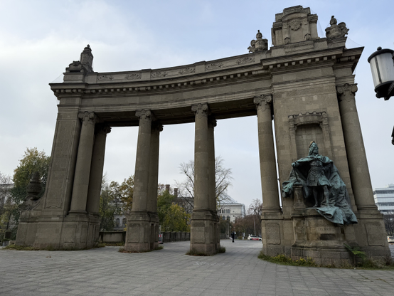 One side of the Charlottenburg tor. It's an imposing stone gate, with a grey statue of a man to the right, and columns keeping the top up.

There are many embellishments carved in the structure itself.

Photo taken in Charlottenburg.