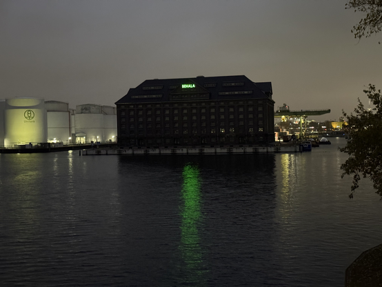 An evening photograph of an old warehouse in a harbour, surrounded by water. The warehouse has a green neon sign saying "Behala". To the left of the warehouse there are silos, and on the right there is a big crane but rather far away. All the lights are reflected in the water.

Picture taken in Moabit.