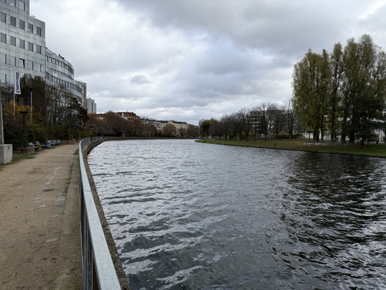 The Spree river, taken from its left shore. On the near shore, there is a gravel path with benches which are in front of wavy glass buildings.

To the right, there is a grassy area with a few trees that are in the process of losing their foliage, and a few grey buildings peaking behind them.

The watter is choppy and mostly reflects the grey sky colour.

Picture taken in Moabit.