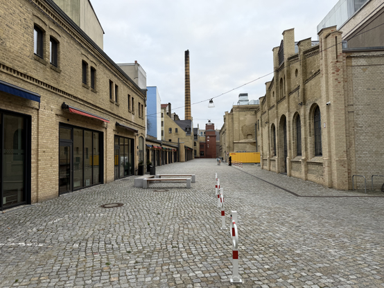 The courtyard of an old factory campus. The ground is paved with cobblestones, and all the buildings are grey/yello brick. There is an old chimney in the distance. Buildings on the left have ground to ceiling windows with doors in then, they also have an  awning above them.

Picture taken in Moabit.