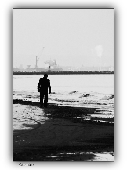 A black and white photograph depicts a man walking along the edge of a beach. The beach has patches of snow scattered around. In the background, there is a port with cranes and industrial structures visible. The man is walking towards the water, and the waves are gently lapping at the shore.