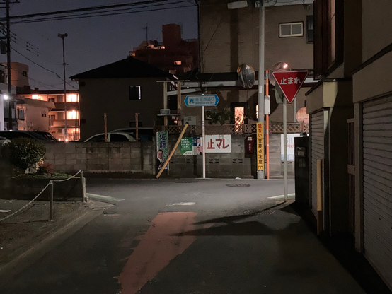 A dimly lit urban street at night, featuring a dead-end sign and various directional signs. There are houses and buildings in the background with illuminated windows. A small tree and a fence are visible on the left side, adding to the residential atmosphere.