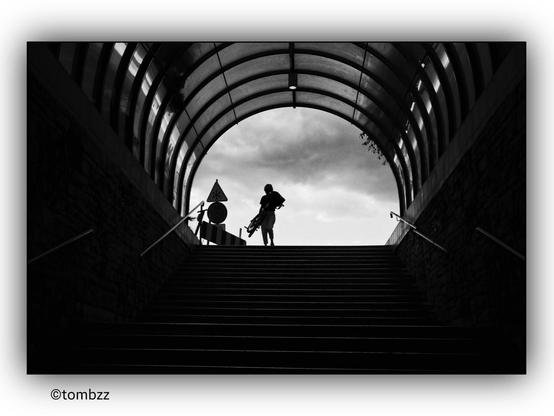 A girl carrying a bicycle is descending a set of stairs into an underpass. The photograph is in black and white. The girl and bicycle are silhouetted against the bright sky, creating a striking contrast. The underpass has a curved, transparent roof with visible structural supports. There are handrails on both sides of the stairs. At the top of the stairs, there are traffic signs and barriers. The stark difference between the dark silhouette and the light background adds a dramatic effect to the image.