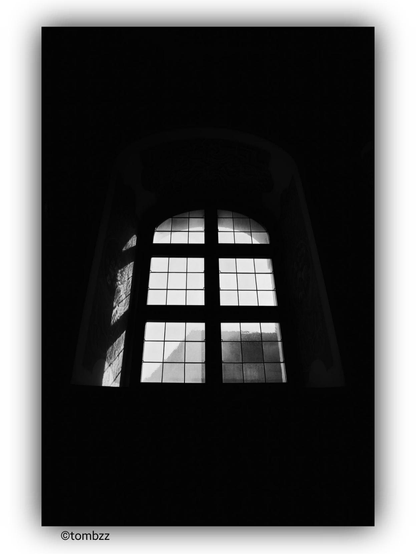 The image depicts a black and white photograph of an arched window inside a church building. The window features a grid of small panes divided by muntins. Bright light streams through the window, creating a high contrast with the dark interior. The light highlights the intricate details of the window frame and the surrounding wall, including decorative elements. Outside the window, part of another building is visible, appearing to be a mountain or a large structure in the background.