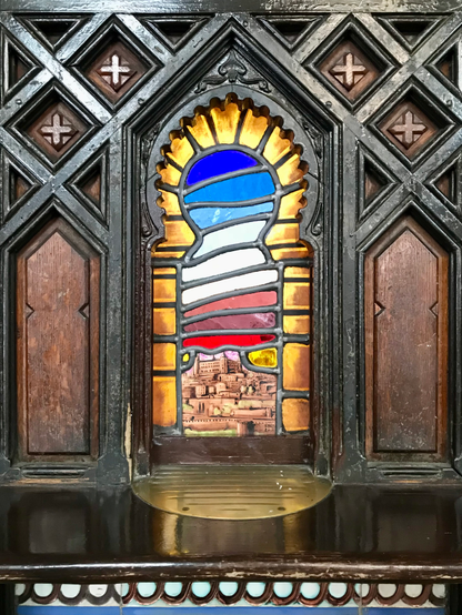 A colorful leaded window depicting the city of Toledo at dawn, with the city at the bottom and layers os sky above in red fading to dark blue to the top. The shape of the window is that of an Arab arch, and the window is surrounded by a panel of aged wood, with veins of deep brown, and adorned with a lattice of diamonds with little crosses inside. 