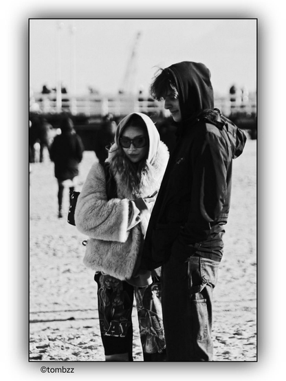 A black and white photograph of a young man and a young woman standing on a beach. The woman is wearing a hooded fur coat and patterned pants, while the man is wearing a hooded jacket and jeans. In the background, there is a blurred view of a pier and several people walking. The image has a soft focus, giving it a nostalgic and candid feel.