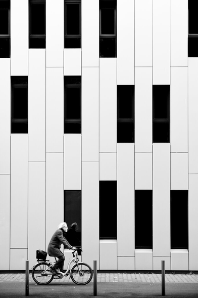 black and white. portrait format. urban scene. a darkly dressed, gray-haired, stylish older man rides a bicycle through the city. the background of the photo is a brightly clad facade with narrow, high windows whose panes are tinted dark. the facade is somehow reminiscent of a piano due to its narrow light and dark elements. the silver-haired head of the man is reflected in one of the dark windows.