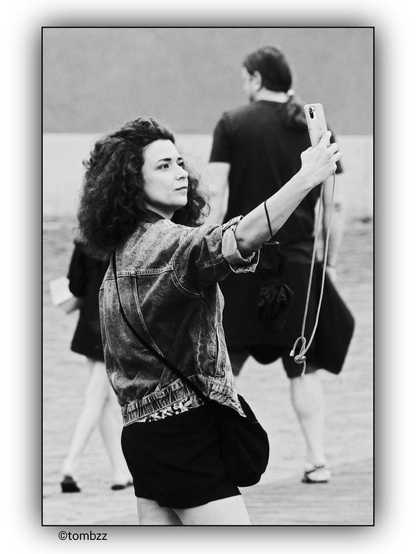 A woman with loose, curly hair takes a selfie. She is wearing a denim jacket and a black skirt, with a black bag slung over her shoulder. In her right hand, she holds a phone, and a lanyard hangs from her wrist. Her face shows concentration, as if she is perfecting the ideal frame. In the background, a man in a dark T-shirt and a girl in light-colored pants are partially visible. The black-and-white photo emphasizes contrasts and the play of light.