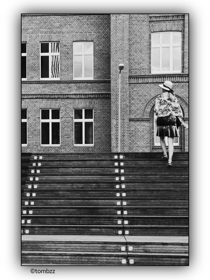 A woman seen from behind is climbing wide wooden stairs with metal connecting elements. In front of her stands a two-story brick building with large, symmetrically arranged windows framed in white. She wears a hat, a floral-patterned blouse, and a skirt adorned with fringes. The black-and-white photo highlights the texture of the bricks and wooden stairs.