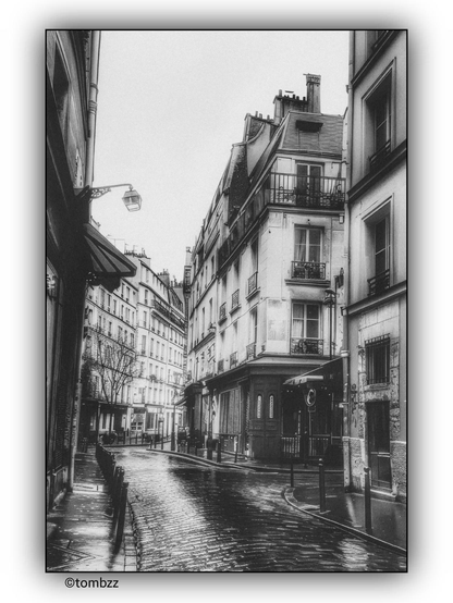 A black-and-white photograph captures a wet street in Paris after the rain. In the foreground, there is a curve in the street, surrounded by characteristic buildings with balconies and tall windows. On the left side, a streetlamp and an awning above the entrance to one of the buildings are visible. The street is separated from the sidewalk by a row of bollards. The black-and-white tones and wet pavement give the photograph a nostalgic, atmospheric feel.