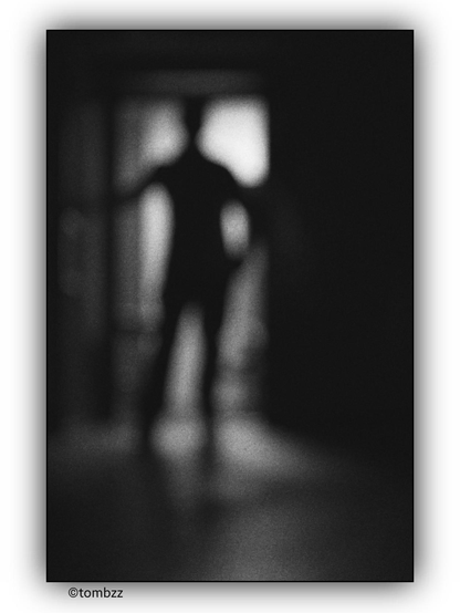 The black-and-white photograph depicts a human silhouette, most likely a man, standing in an open doorway. Light from outside sharply cuts through the darkness of the interior, casting a contrasting glow on the figure, blurring its contours. The grainy texture of the image adds rawness and mystery, emphasizing an atmosphere of ambiguity. Is the person entering, leaving, or lingering in hesitation? The shadowy room remains concealed, while the brightness of the doorway seems to be the only window to the outside. The photograph raises more questions than answers, inviting interpretation.