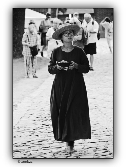 The black-and-white photograph captures a young woman walking barefoot on a cobblestone street. Her outfit draws attention—a stylishly cut dress flows gently around her legs, while a large straw hat shields her face, adding an air of mystery. She wears glasses, accentuating her intriguing presence. In her hands, she carries a small bowl of food, moving forward calmly and confidently.

The background brings the scene to life—in the distance, a group of people is engaged in their activities, some walking in the same direction as the woman, others deep in conversation. The street, the cobblestones, and the surrounding people provide context, creating an image of a vibrant place where the central figure stands out with her presence and style.